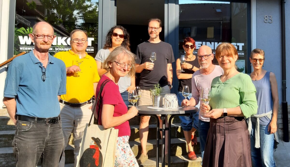 Ein Gruppenfoto von neun Menschen, die in die Kamera schauen. In ihren Händen Weingläser.