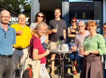 Ein Gruppenfoto von neun Menschen, die in die Kamera schauen. In ihren Händen Weingläser.