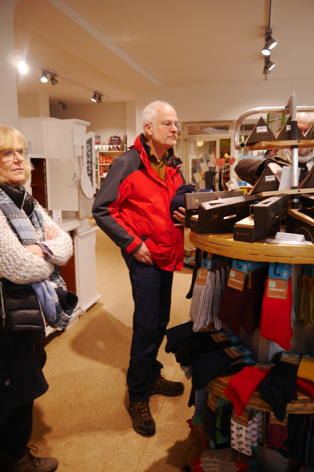 Mann mit roter Jacke steht vor einem drehenden Regal und sieht sich dessen Inhalt - Socken - an. 