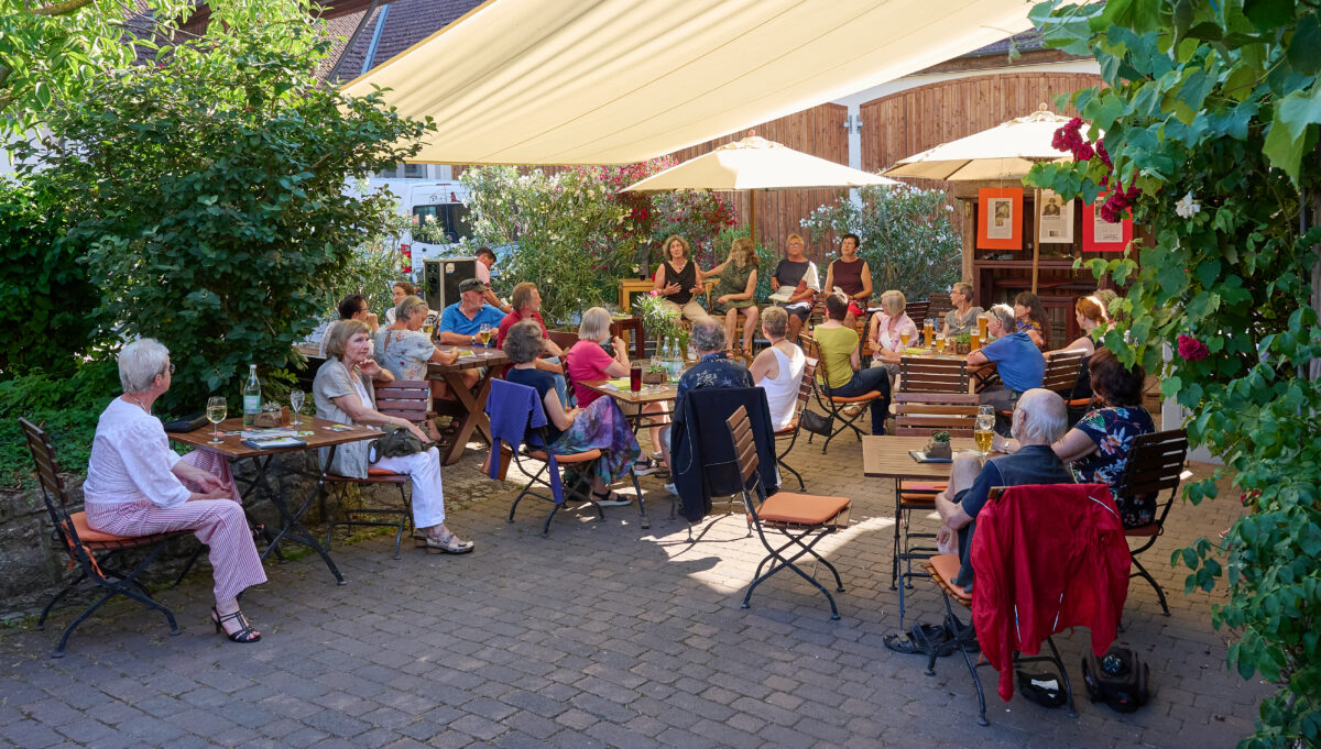 Foto vom Innenhof des Cafés. Die Besucher*innen sitzen an Tischen und lauschen den Worten der vier(ehemaligen) Politikerinnen. 