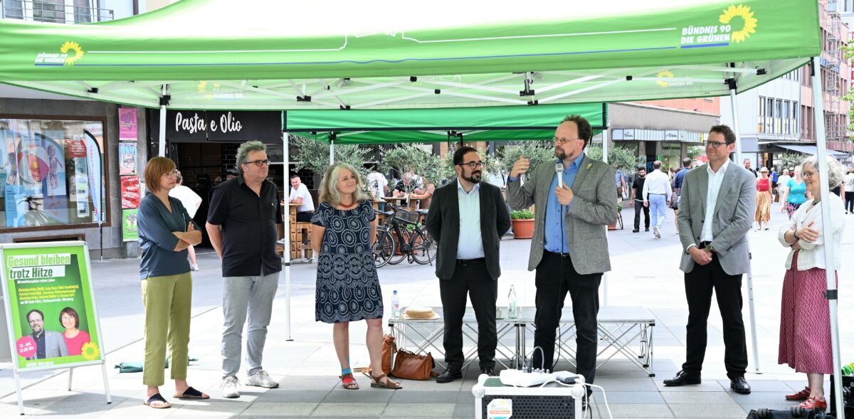 Die im Text erwähnten Politiker und Politikerinnen unter einem grünen Pavillon in der Würzburger Innenstadt, genauer am QR-Platz. Patrick Friedl hat ein Mikrofon in der Hand und erklärt etwas, die Anderen hören ihm zu und sehen ihn dabei an. Im Hintergrund ist ein Plakat mit Flyer der Veranstaltung und Passanten zu sehen. 