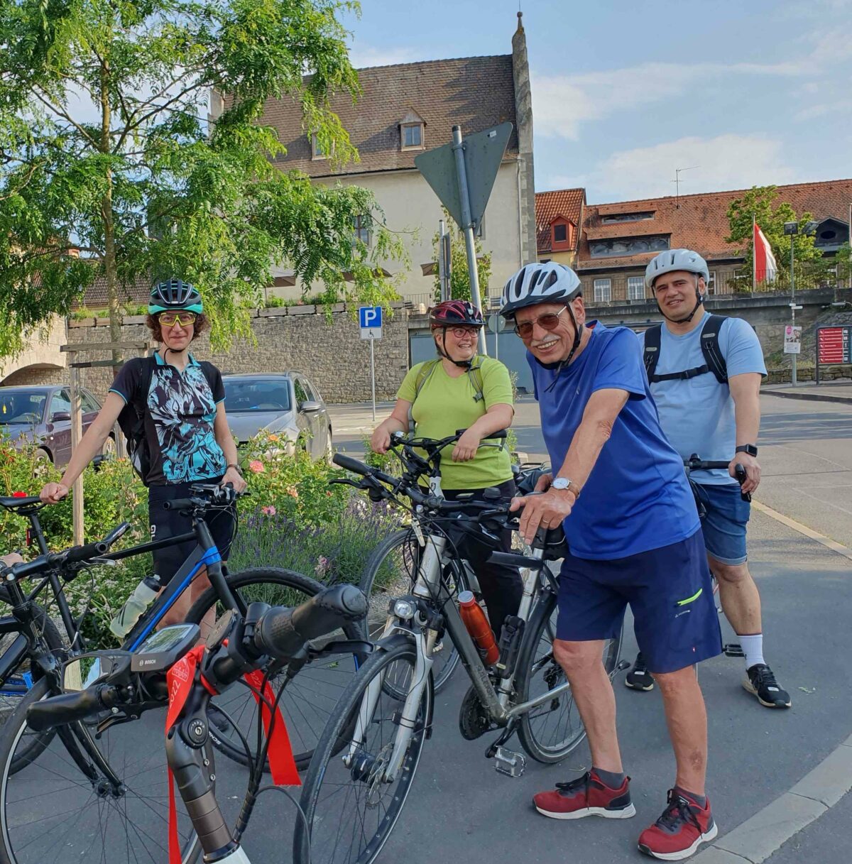 Zwei Frauen und zwei Männer der Radtruppe lächeln in die Kamera, während sie eine Pause zu machen scheinen. Im Hintergrund eine Dorfstraße. 