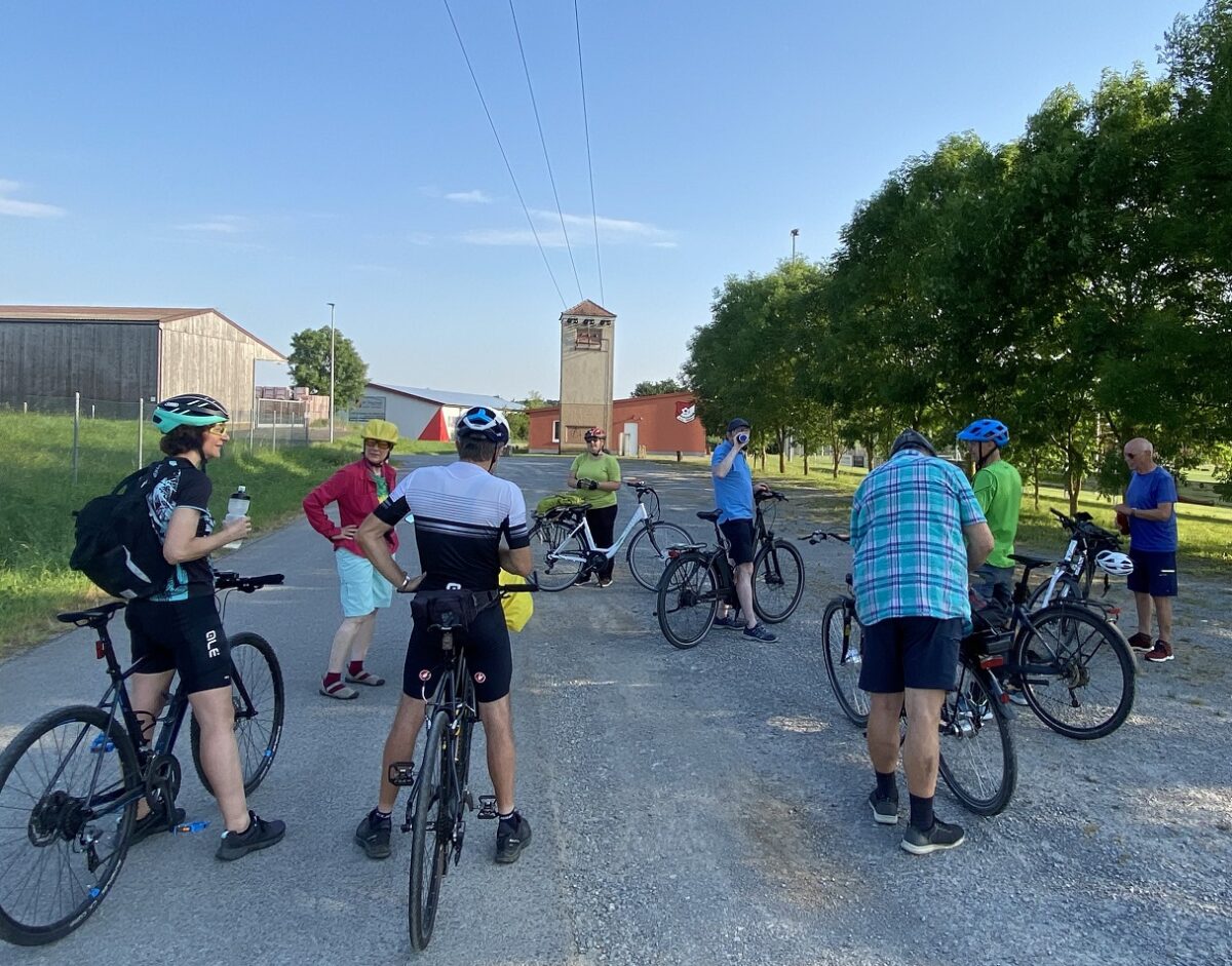 Zu Sehen ist die Gruppe der Fahrradfahrenden von hinten- es scheint alles machen sie eine Pause. Jeder trägt einen Helm und sommerliche Kleidung. Im Hintergrund ist ein blauer Himmel und grüne Bäume zu sehen, sowie ein Turm und Häuser, die ein Dorf andeuten. 