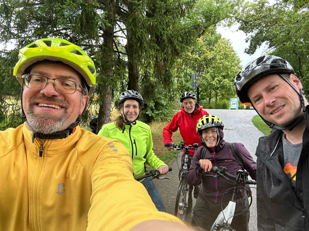 Selfie von fünf Menschen, davon drei Männer und zwei Frauen. Alle sind mit Fahrradhelm und Fahrrädern, sowie Regenjacken ausgestattet. Im Hintergrund ist ein Fahrradweg und grüne Bäume zu sehen. Das Wetter scheint regnerisch zu sein, der Himmel ist grau.