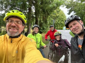 Selfie von fünf Menschen, davon drei Männer und zwei Frauen. Alle sind mit Fahrradhelm und Fahrrädern, sowie Regenjacken ausgestattet. Im Hintergrund ist ein Fahrradweg und grüne Bäume zu sehen. Das Wetter scheint regnerisch zu sein, der Himmel ist grau.