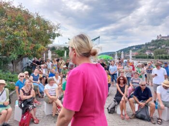 Foto von der Veranstaltung, aus einem Winkel in dem Katharina Schulze im rosa Kleid von hinten zu sehen ist und mit Blick in die Kamera das Publikum. Das Publikum sitz auf weisen Hockern oder steht, ist sommerlich gekleidet und hört Katharina Schulze zu.