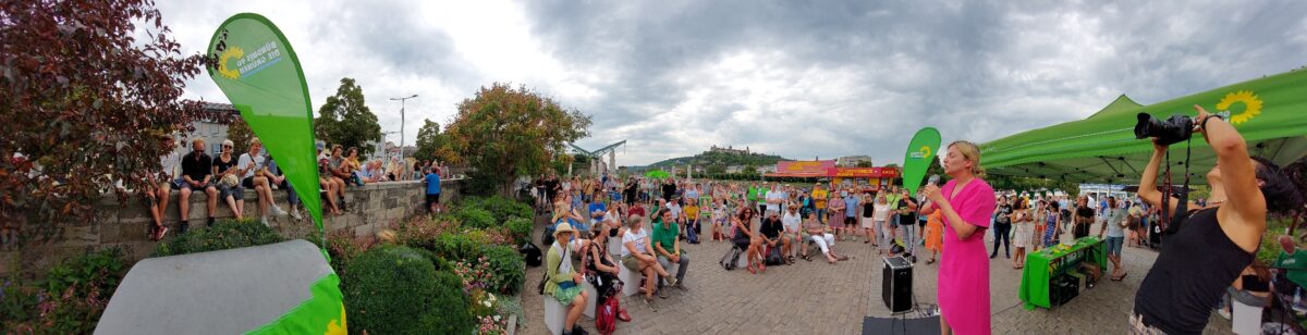 Panorama Bild von der Veranstaltung "Auf ein Eis mit Katha". Rechts im Bild eine Fotografin mit gehobener Kamera, daneben Katharina Schulze mit Mikrofon in der Hand, im restlichen Teil des Bilder Publikum zu sehen und ein bewölkter Himmel
