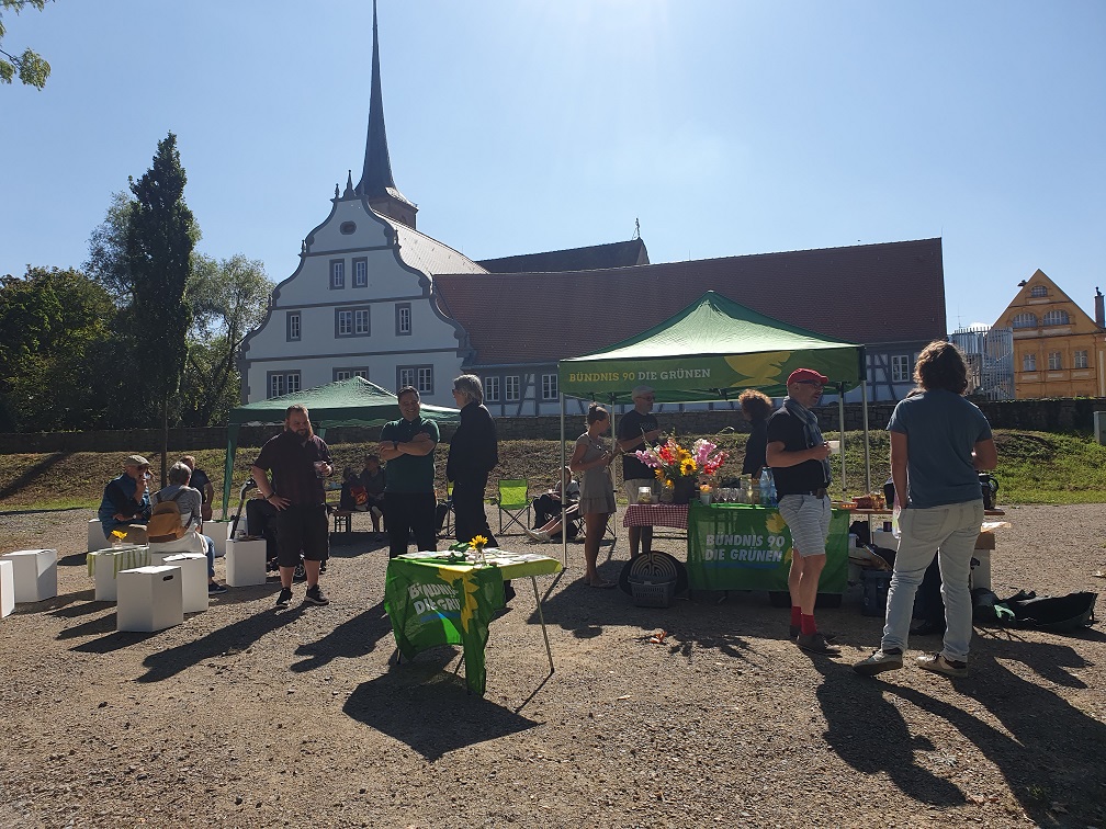 Foto mit Tischen und Stühlen. Auf den Stühlen sitzen Menschen und unterhalten sich. Im Hintergrund ist eine Kirche zu sehen. Auf einem Tisch ist der Brunch und Blumen zu sehen.