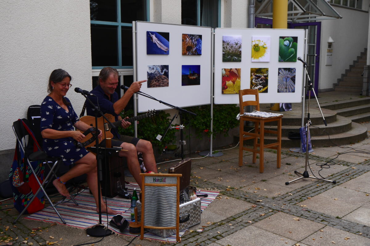 Links im Bild eine Frau mit einer Art Rassel in der Hand, daneben ein Mann mit Gitarre. Beide sitzen auf Stühlen vor Mikrofonen. Rechts im Bild zwei Aufstellwände mit bunten Fotografien. Davor ein Stuhl und ein Mikrofon, hier hat vermutlich die Lesung stattgefunden. 