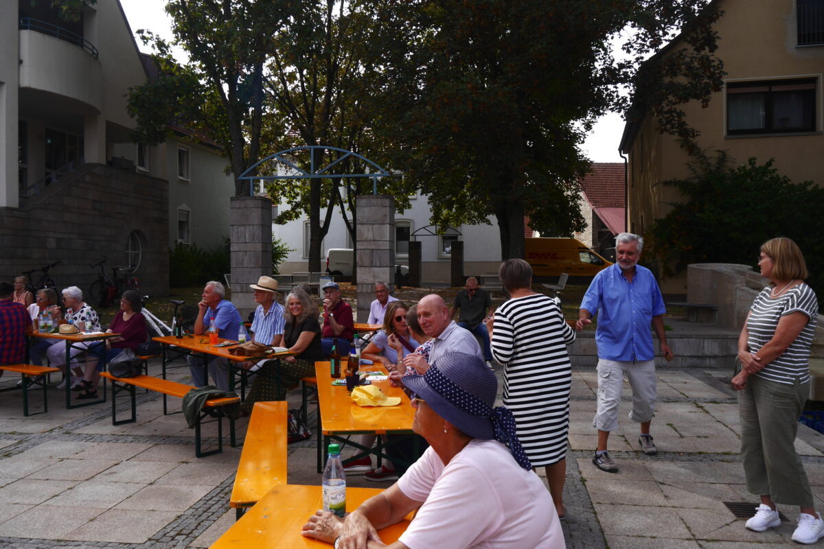 Auf orangenen Bierbänken sitzen Menschen in Sommerkleidung, im Hintergrund sind in unmittelbarer Nähe Bäume und Häuser zu sehen.