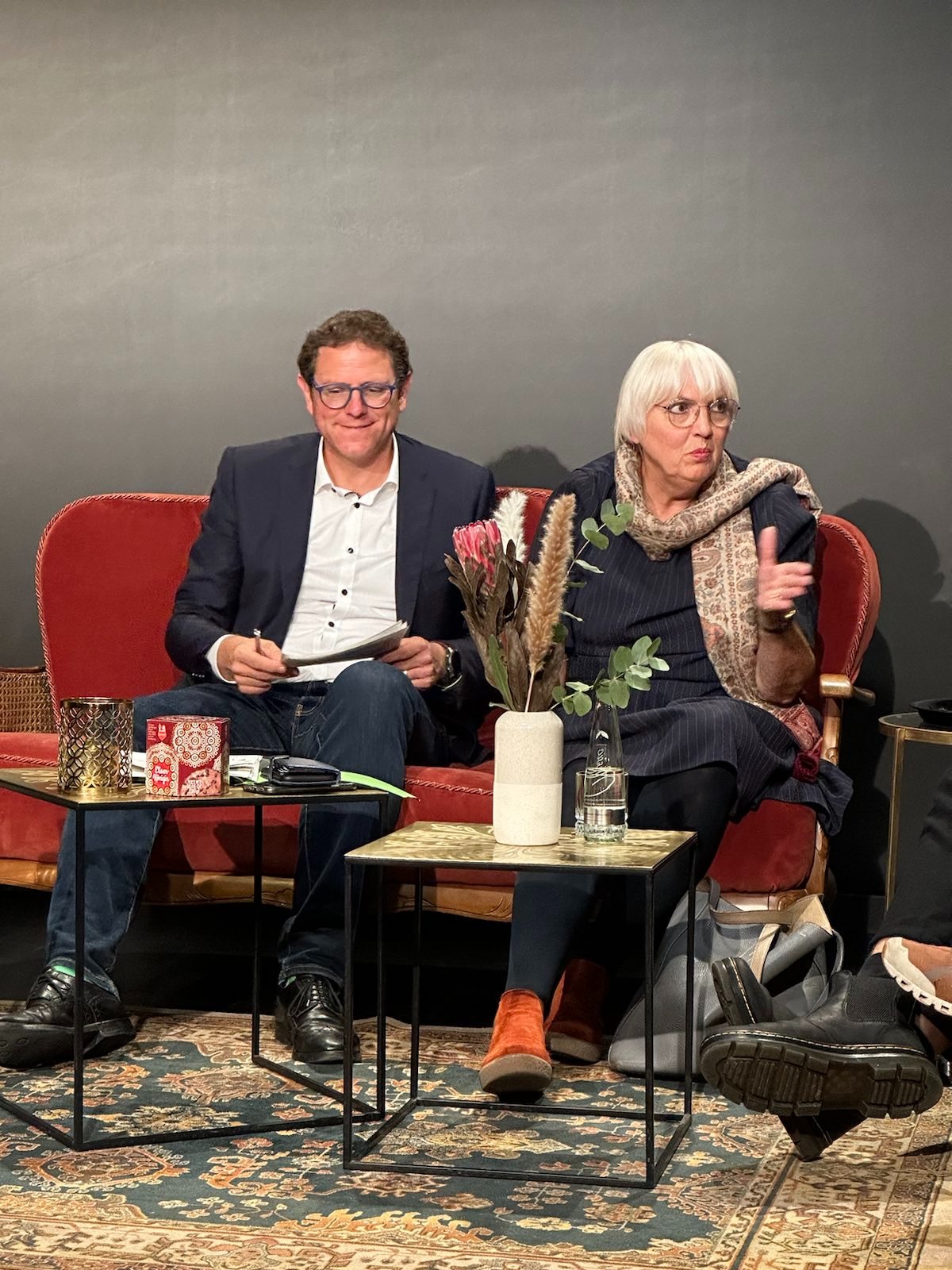 Ein Foto von Claudia Roth und Martin Heilig auf einen roten Sofa. Vor ihnen zwei kleine Tische mit Blumen und Notizen darauf.