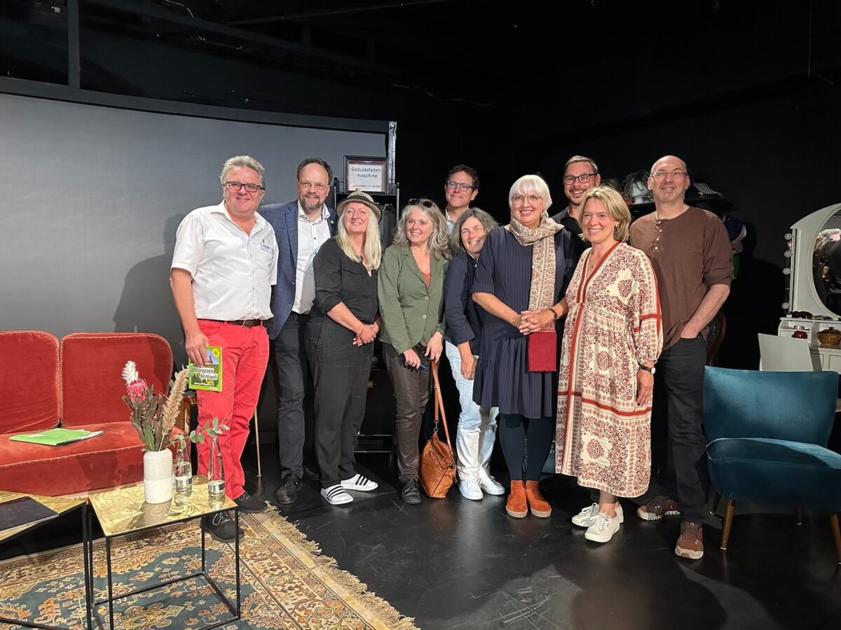 Ein Foto mit den Teilnehmenden der Podiumsdiskussion auf der Bühne. Links neben den Personen steht ein rotes Sofa mit einen kleinen Tisch.