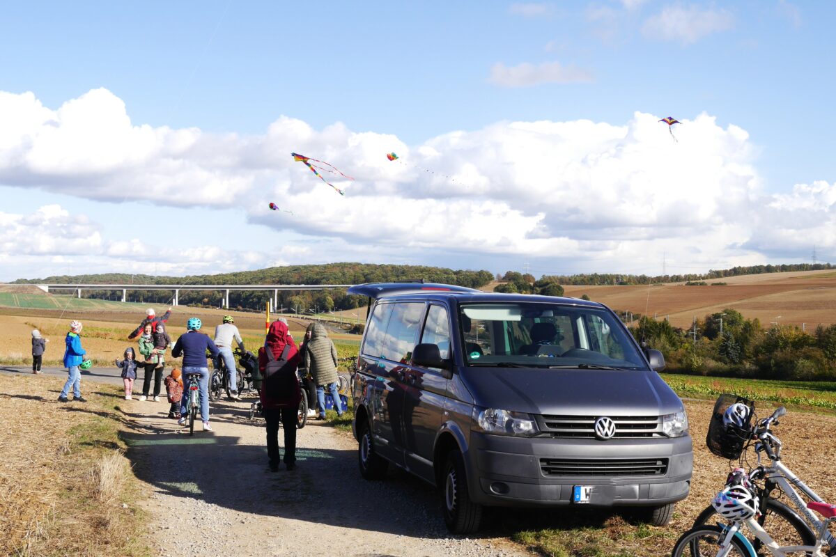 Um einen VW Bus herum, der dort geparkt ist, tummeln sich allerlei Menschen herum und bestaunen das Drachensteigen