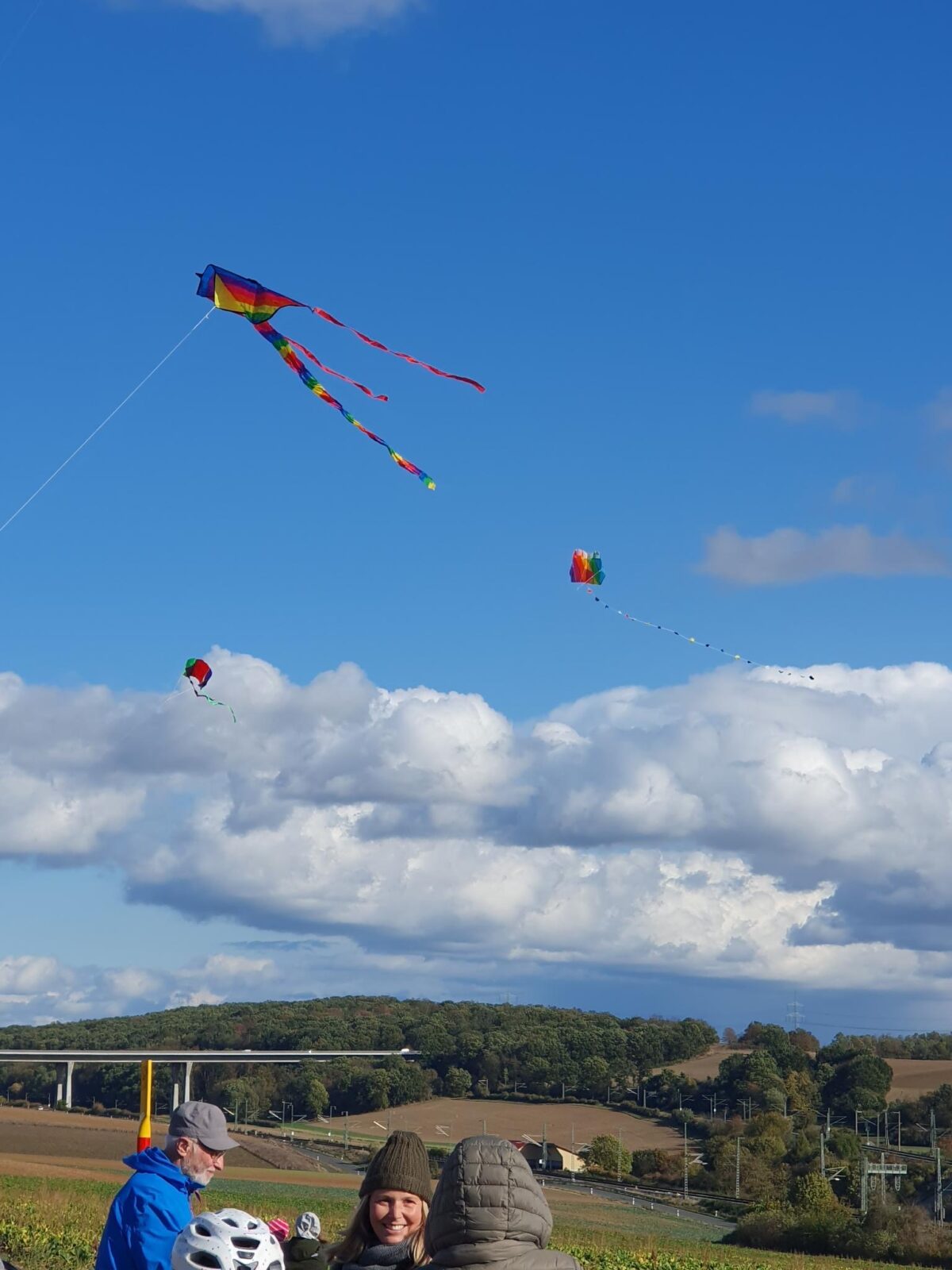 Drei Drachen wehen im Wind, im unteren Teil des Bildes drei Menschen in warmer Kleidung zu sehen