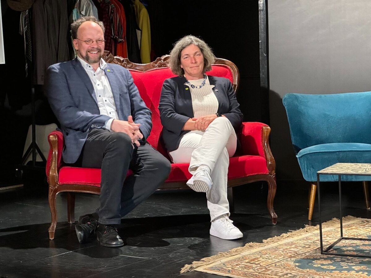 Ein Foto von Patrick Friedl und Kerstin Celina auf einem roten Sofa. Rechts daneben ein blaues Sofa und ein kleiner Tisch. Das Foto wurde auf der Bühne gemacht