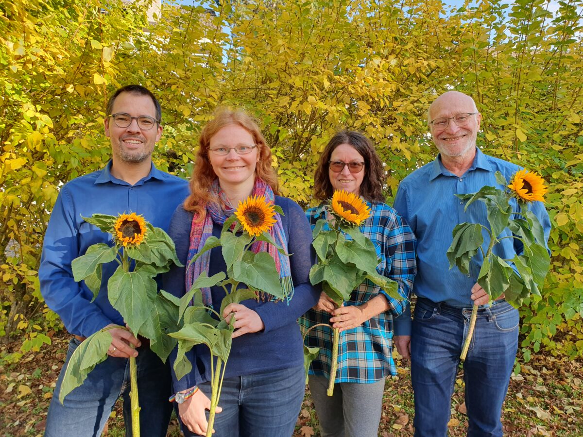 Die vier Mitglieder des Vorstandes stehen mit Sonnenblumen in der Hand vor gelben Sträuchern und lächeln in die Kamera