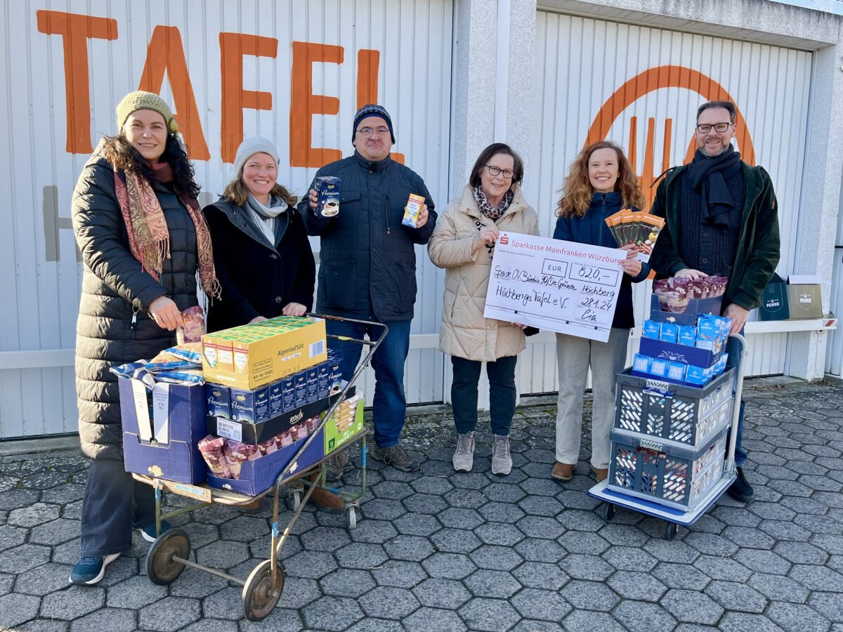 Vertreter*innen des Grünen Ortsverband Höchbergs und die Vorsitzende der Höchberger Tafel stehen gemeinsam vor Garagen mit der Aufschrift "Tafel" in orange. In ihren Händen haben sie einen Scheck über 820€ und diverse Lebensmittel auf kleinen Wägen