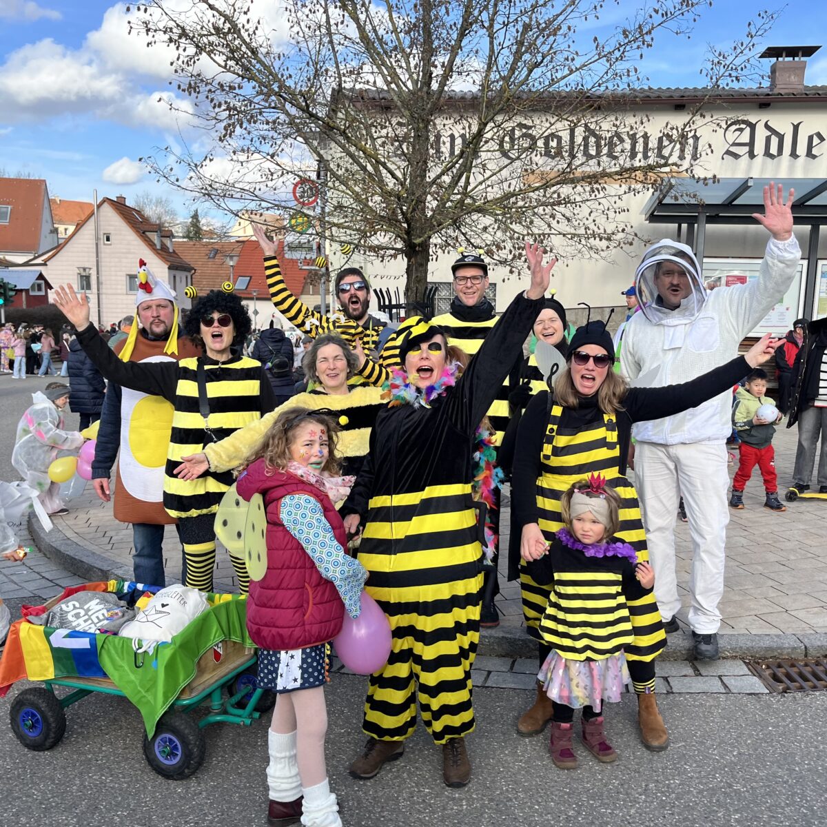 Neun Erwachsene und zwei Kinder verkleidet als Bienen und ein Mann als Imker lächeln mit gestreckten Armen in die Kamera. Sie rufen vermutlich "Helau" und im Hintergrund ist ein Faschingsumzug zu erkennen. Strahlend blauer Himmel deutet auf gutes Wetter hin. 