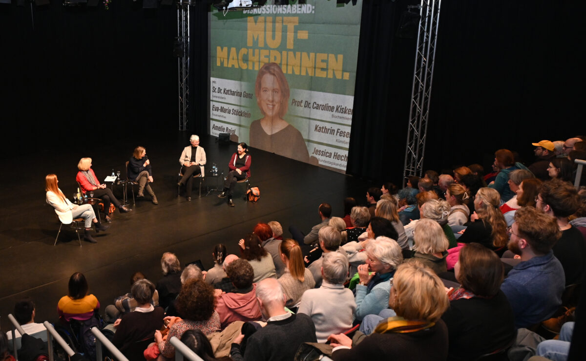 Das Bild ist von oben fotografiert. Man kann das Publikum erkennen und die Bühne auf der die Diskussionsteilnehmerinnen sitzen.