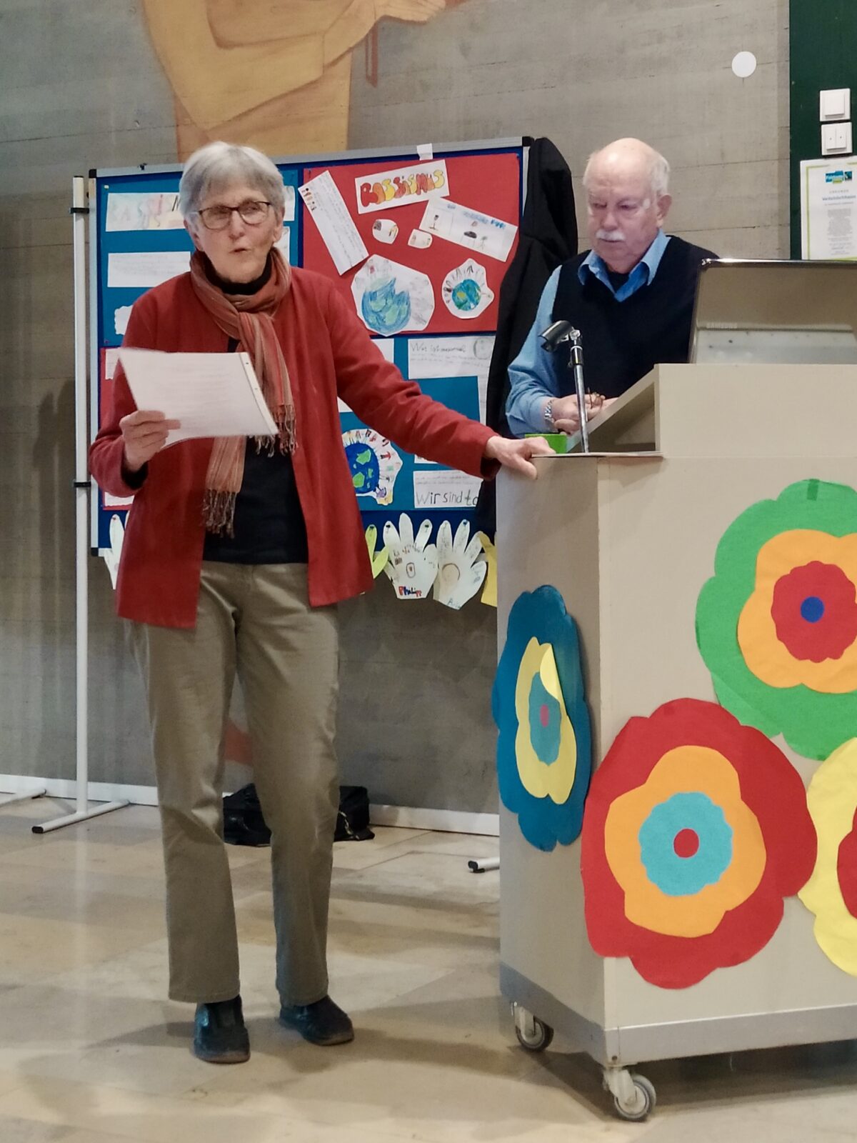 Ärzteehepaar Frau Dr. Päivi Köhler und Herr Dr. Bernhard Köhler aus Randersacker zu sehen. Frau Köhler beim Vortrag am Stehpult, das mit bunten Papierblumen geschmückt ist.