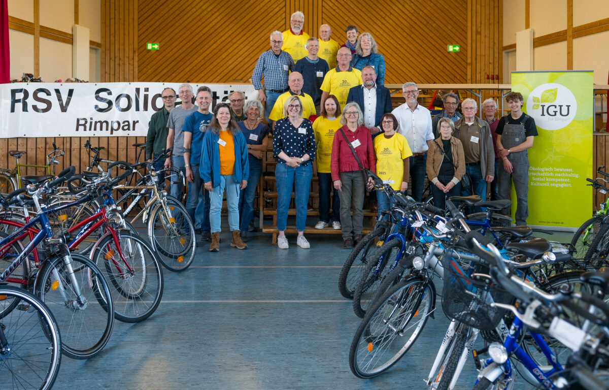 Die 24 Helfer*innen des Teams Fahrradflohmarkt stehen für ein Gruppenfoto hinter den rechts und links in der Halle aufgestellten Fahrrädern.